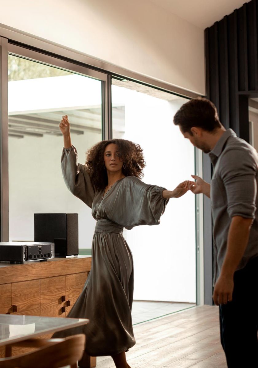 A couple dancing in a modern living room with an audio system on a wooden cabinet nearby.