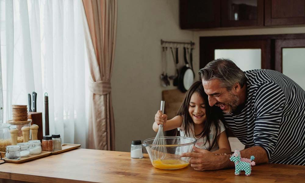 A father and daughter laughing while whisking ingredients together in a bright kitchen.